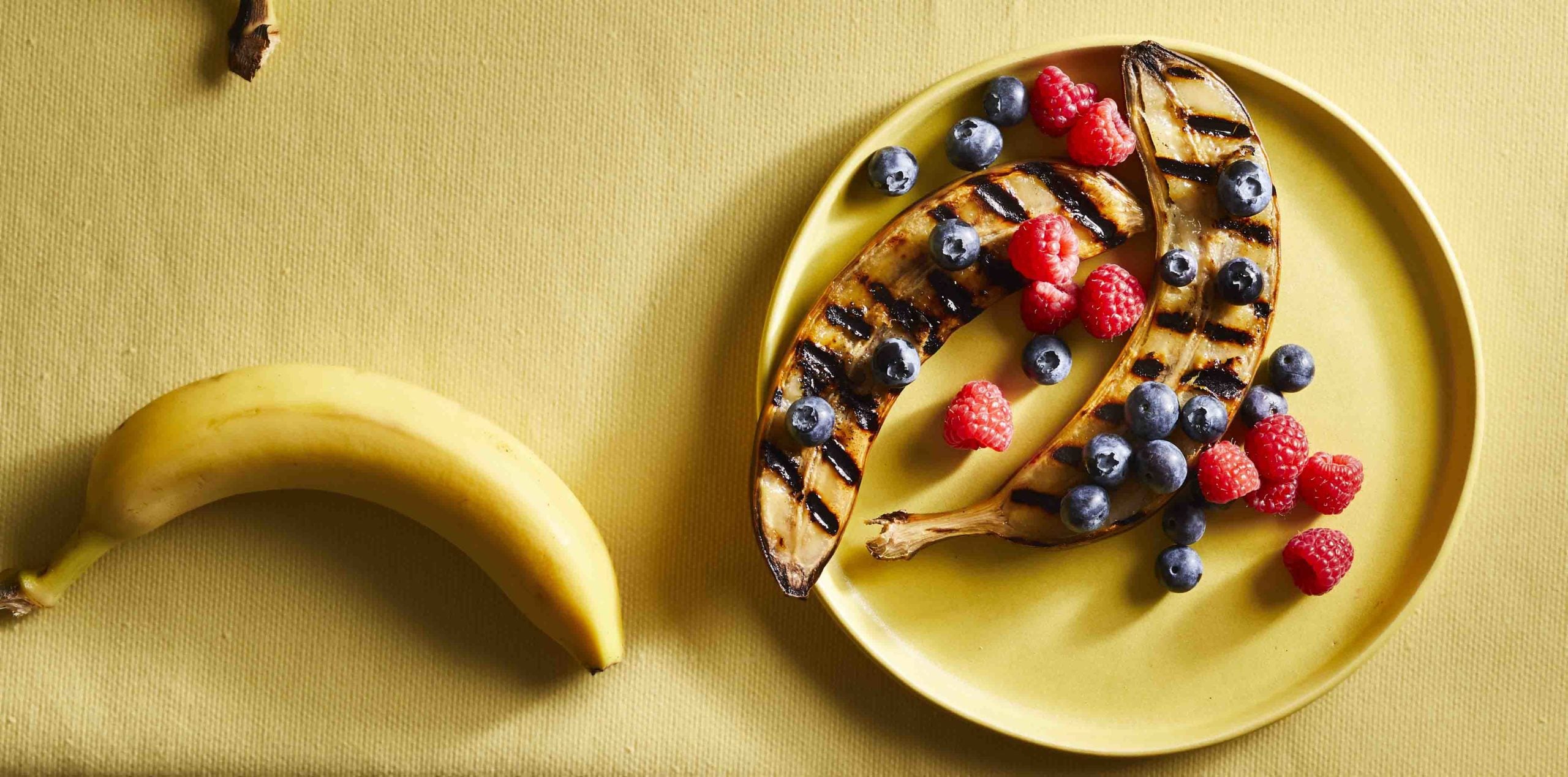 Bananas with peel on next to a plate of peeled grilled bananas topped with berries on a yellow plate, all sitting on top of a yellow tablecloth 